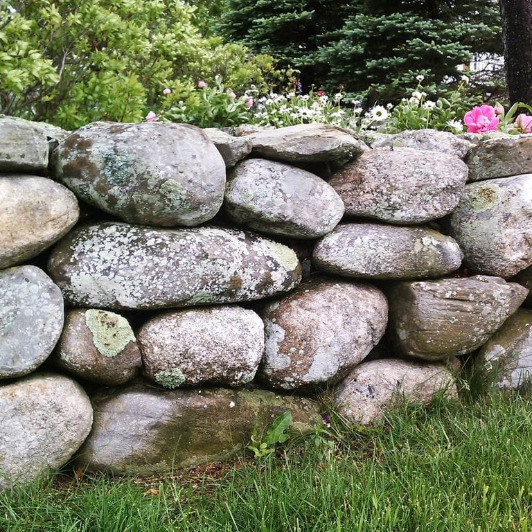 Feature image for New England Fieldstone Wallstone - Rounds. The image shows this product being used in a landscape wallstone application. The dominant color shown is gray; with beige, black, brown, green, orange/rust, pink, purple, white, tan and cream secondaries. Random and speckled color patterns.