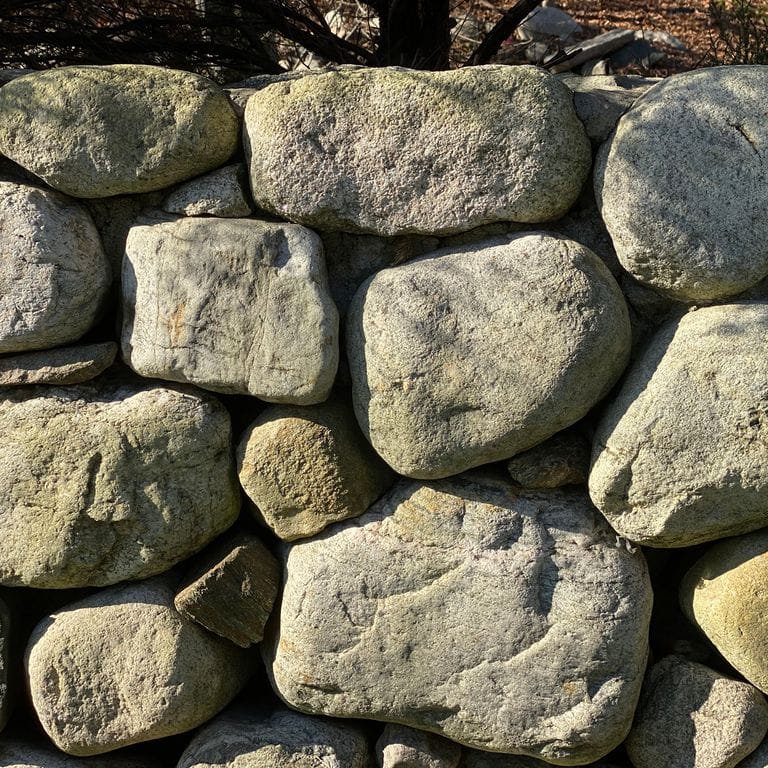 Best close-up image for New England Fieldstone - Buff Rounds Wallstone. The image shows this product being used in a landscape wallstone application. The surface finish shown is weathered. Dominant colors shown are beige, brown, gray and tan; with green, orange/rust, yellow and cream secondaries. The color mix creates a warm feeling. Regular color pattern.