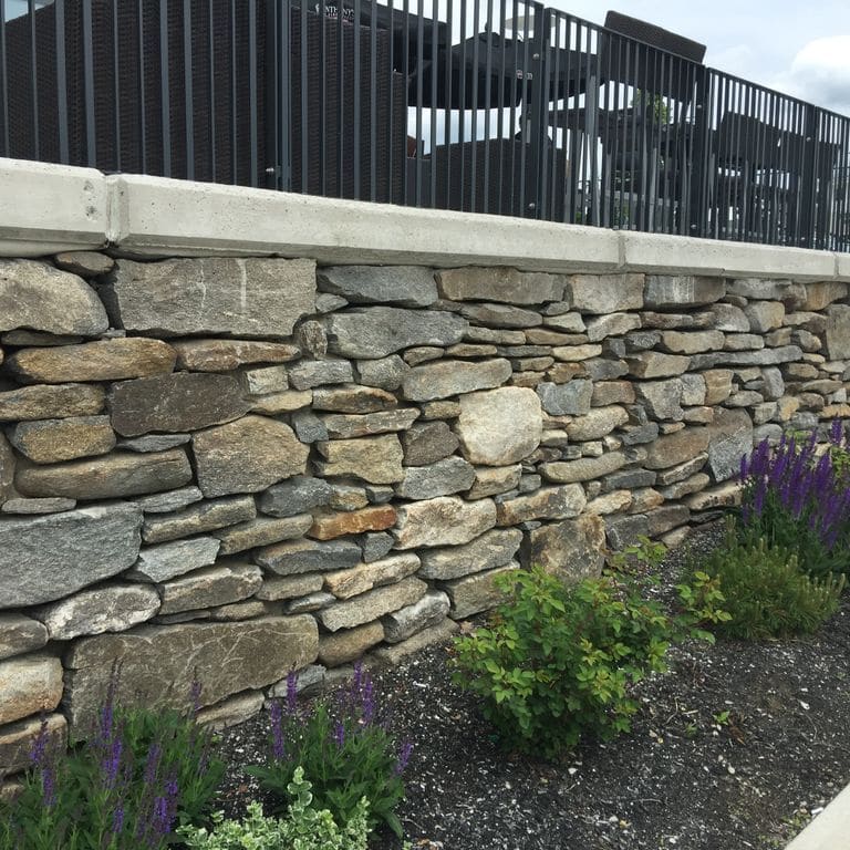 Feature image for New England Fieldstone Wallstone - Barre Dark. The image shows this product being used in a landscape wallstone application. The design pattern shown is ledgestone. Dominant colors shown are beige, brown and gray; with orange/rust, purple, white, tan and cream secondaries. The color mix creates a warm feeling. Random, speckled and cloudy color patterns.