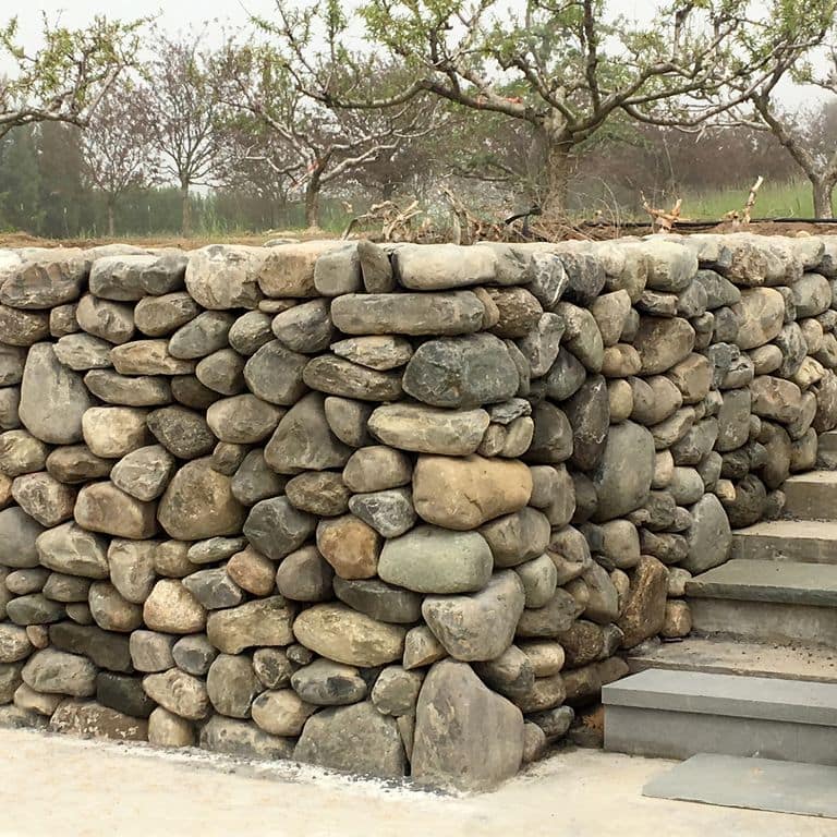 Feature image for New England Fieldstone Wallstone - Medomak Gray Rounds. The image shows this product being used in a landscape wallstone application. The design pattern shown is mixed medium rounds. Dominant colors shown are beige, brown, gray and tan; with green, orange/rust, purple, white and cream secondaries. The color mix creates a warm feeling. Random color pattern.