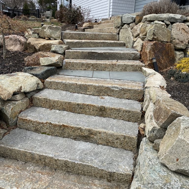 Application image for Reclaimed First Generation Granite Curbstone Steps - Textured Riser and Tread. The image shows this product being used in a monolithic steps application. This is a landscape stone for outdoor commercial and residential applications. Suitable for general use, freeze-thaw conditions, low heat absorption surfaces, high traffic areas and low slipperiness surfaces situations. Higher to high on the price spectrum.