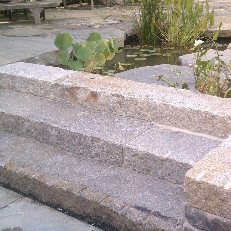 Application image for Reclaimed Second Generation Granite Curbstone Steps - Split Face Riser. The image shows this product being used in a monolithic steps application. This is a landscape stone for outdoor commercial and residential applications. Suitable for general use, freeze-thaw conditions, low heat absorption surfaces, high traffic areas and low slipperiness surfaces situations. Higher to high on the price spectrum.