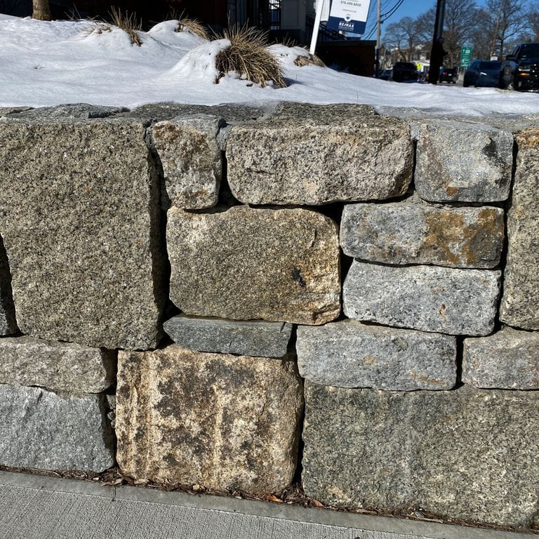 Application image for Reclaimed Granite Block Wallstone - Split Block Mix. The image shows this product being used in a landscape wallstone application. The surface finish shown is a combination of split-face and weathered. The dominant color shown is gray; with beige, brown, green and tan secondaries. The color mix creates a warm feeling. Speckled color pattern.