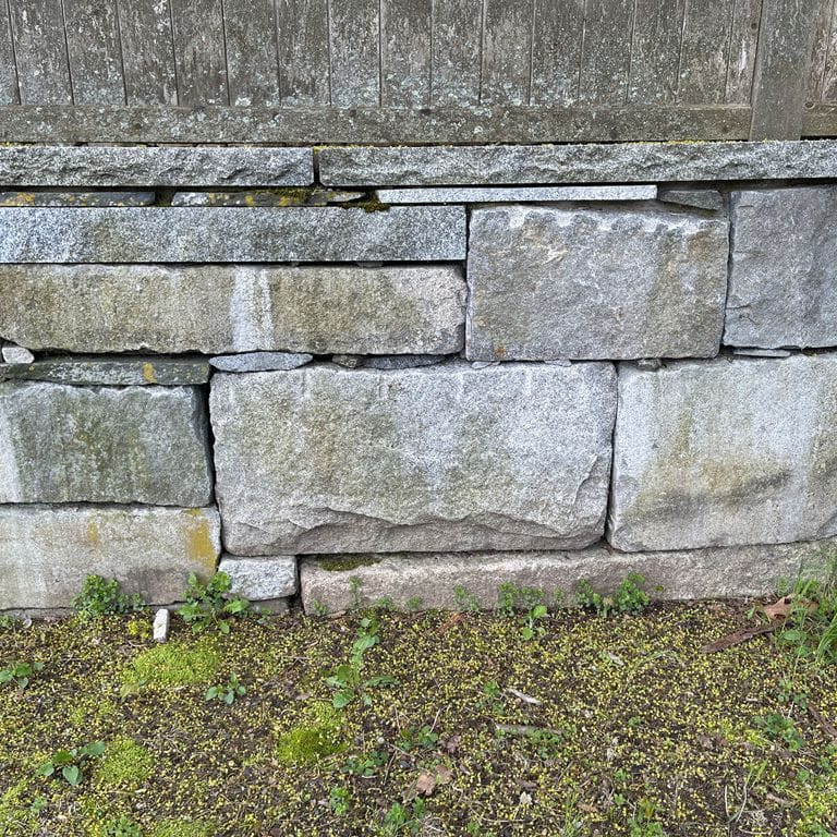 Application image for Reclaimed Granite Block Wallstone - Split Block Mix. The image shows this product being used in a landscape wallstone application. The surface finish shown is a combination of split-face and weathered. The dominant color shown is gray; with blue, brown, green and white secondaries. The color mix creates a cool feeling.