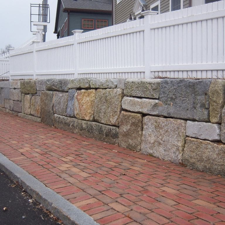 Feature image for Reclaimed Granite Block Wallstone - Block Mix. The image shows this product being used in a landscape wallstone application. The design pattern shown is quarried-snapped roughly square roughly rectangular with sharp edges. Dominant colors shown are brown and gray; with beige, black, blue, green, orange/rust, pink, purple, white and tan secondaries. The color mix creates a warm feeling. Speckled, blotched and cloudy color patterns.