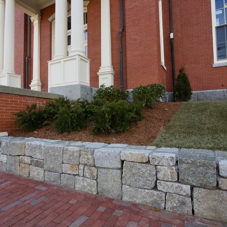 Application image for Reclaimed Granite Block - Wallstone. The image shows this product being used in a landscape wallstone application. The dominant color shown is gray; with brown, orange/rust, purple, white and tan secondaries. Speckled and blotched color patterns.