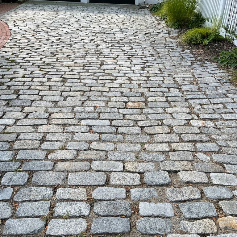 Application image for Reclaimed New England Granite Cobblestones - Medium Pavers. The image shows this product being used in a driveway pavers application. The surface finish shown is a combination of weathered and worn. The dominant color shown is gray; with black, brown and white secondaries. The color mix creates a cool feeling. Speckled color pattern.