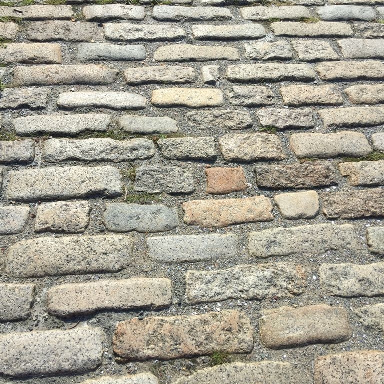 Application image for Reclaimed New England Cobblestones - Jumbo Worn Top. The image shows this product being used for driveway pavers and path pavers. Dominant colors shown are beige and gray; with brown, green, white, tan and cream secondaries. The color mix creates a warm feeling. Speckled and blotched color patterns.