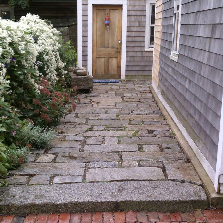 Feature image for Reclaimed New England Granite Cobblestones - Super Jumbo Pavers. The image shows this product being used in a path pavers application. Design patterns shown are random with wide sides up and random with tops up. The dominant color shown is gray; with blue, brown, purple and tan secondaries. Speckled and blotched color patterns.