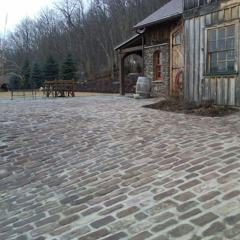 Feature image for Reclaimed Midwest Medina Sandstone Cobblestones. The image shows this product being used for driveway pavers and patio pavers. The design pattern shown is running bond with tops up. The dominant color shown is gray; with beige, brown, green, purple and white secondaries. Speckled and blotched color patterns.