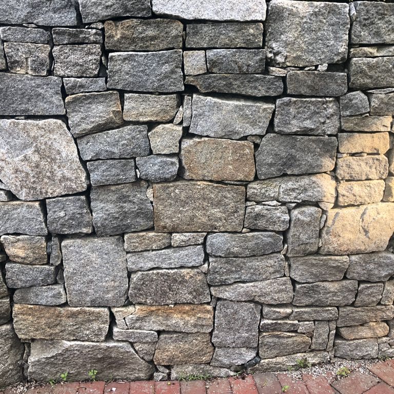 Best close-up image for Granite - Reclaimed New England Quarry Tailings - Wallstone. The image shows this product being used in a landscape wallstone application. The surface finish shown is split-face with flat plane. The dominant color shown is gray; with black, brown, green, orange/rust, white and cream secondaries. The color mix creates a warm feeling. Regular, fine speckled, swirled, speckled, blotched and cloudy color patterns.