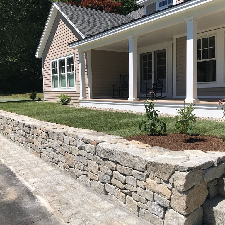 Feature image for Reclaimed New England Granite Quarry Tailings - Wallstone. The image shows this product being used in a landscape wallstone application. The design pattern shown is quarried-snapped mosaic. The dominant color shown is gray; with beige, brown, white, tan and cream secondaries. The color mix creates a warm feeling. Speckled, blotched and cloudy color patterns.