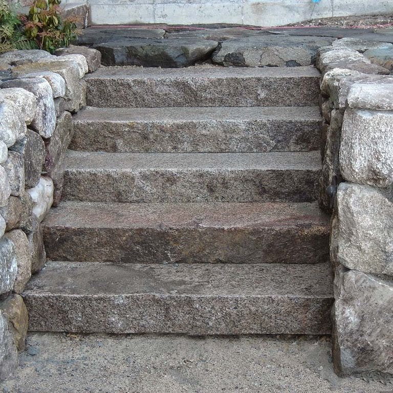 Close-up image for Reclaimed Granite Step - Live Edge. The surface finish shown is flamed. The dominant color shown is gray; with brown, green and purple secondaries. Exhibits "movement". Random, speckled, coarse speckled, blotched, swirled and cloudy color patterns.