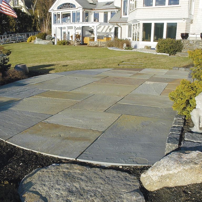 Feature image for Full Color Bluestone - Natural Cleft Pavers. The image shows this product being used in a patio pavers application. The surface finish shown is natural cleft. Dominant colors shown are blue, green and gray; with brown, orange/rust and tan secondaries. Exhibits "movement". Random color pattern.