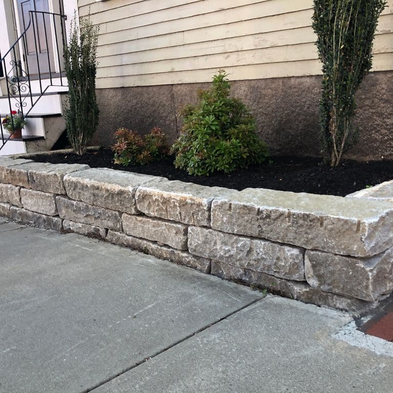 Feature image for Reclaimed Granite Curbstone - Junior Wallstone. The image shows this product being used in a landscape wallstone application. The dominant color shown is gray; with beige, brown, green, purple, white and tan secondaries. Exhibits "movement". Regular, speckled, blotched and cloudy color patterns.