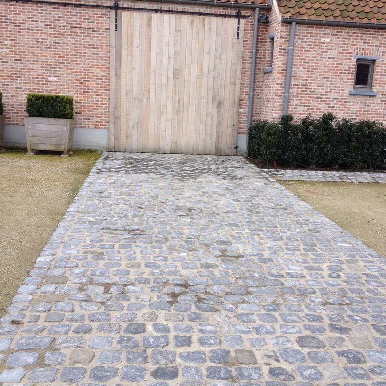 Feature image for Reclaimed Belgian Bluestone Cobblestones - Worn Surface. The image shows this product being used in a driveway pavers application. The surface finish shown is . The dominant color shown is gray; with blue, white and tan secondaries. Exhibits "movement" and the color mix creates a cool feeling. Speckled, blotched and cloudy color patterns.