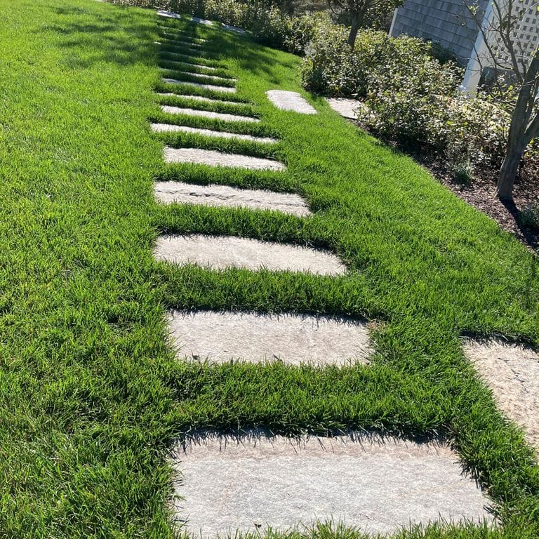 Application image for Reclaimed First Generation Granite Curbstone - Steppers. The image shows this product being used in a path pavers application. The surface finish shown is a combination of weathered and worn. The dominant color shown is gray; with a tan secondary. The color mix creates a warm feeling. Speckled color pattern.