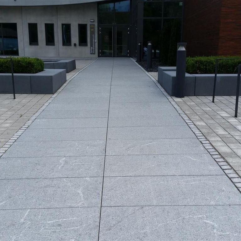 Feature image for Granite - Republic Black - Pavers. The image shows this product being used in a path pavers application. Dominant colors shown are blue and gray; with a white secondary. Exhibits "movement" and the color mix creates a cool feeling. Speckled and cloudy color patterns.