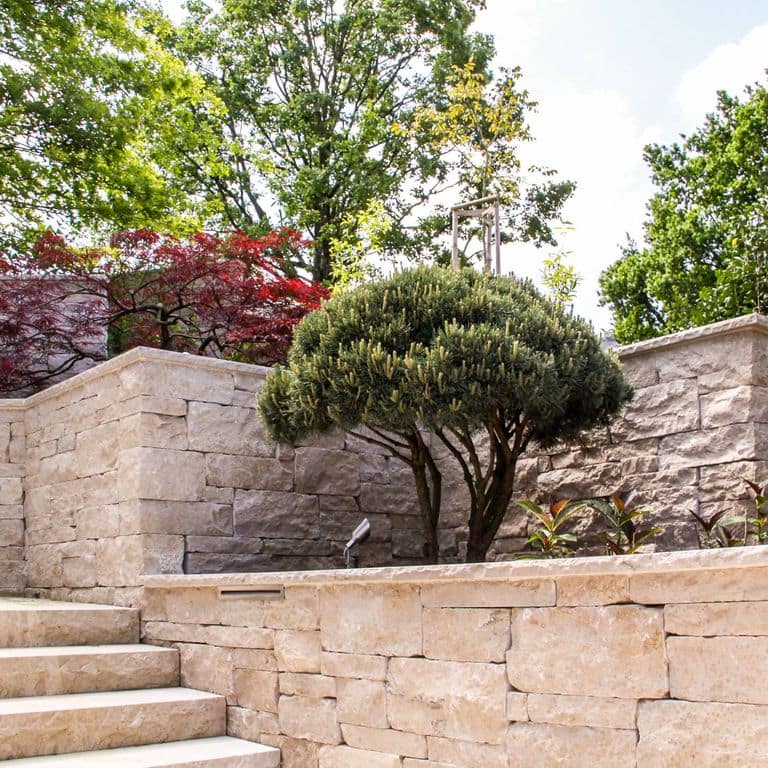 Feature image for Bavarian Limestone - Beige- Wallstone. The image shows this product being used in a landscape wallstone application. Dominant colors shown are beige and tan; with orange/rust, pink and cream secondaries. Exhibits "movement" and the color mix creates a warm feeling. Swirled and cloudy color patterns. Contains mineral inclusions.