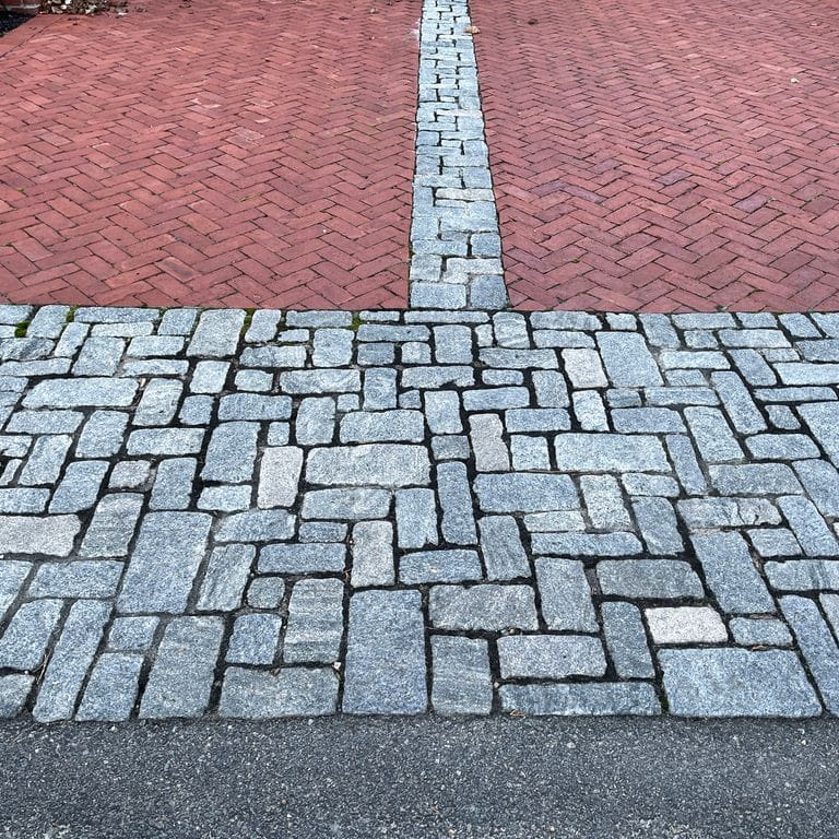 Application image for Granite Cobblestones - Gray. The image shows this product being used for driveway pavers and driveway aprons. The surface finish shown is tumbled. The dominant color shown is gray; with blue and white secondaries. The color mix creates a cool feeling. Streaked and speckled color patterns. Contains some mica and mineral inclusions.