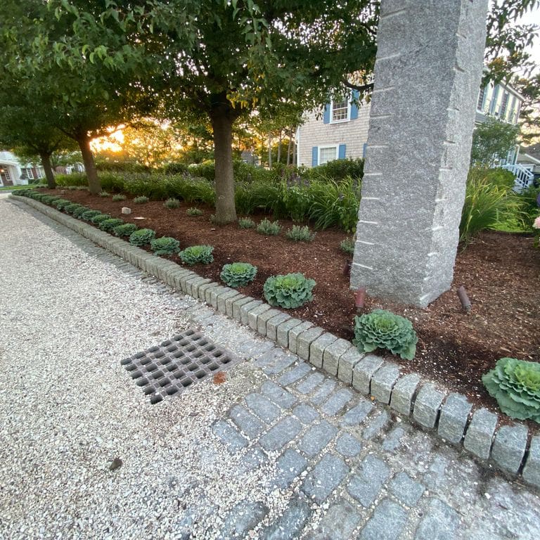 Application image for Gray Granite Cobblestone - Rectangles. The image shows this product being used in a landscape curbing &amp; edging application. The surface finish shown is tumbled. The dominant color shown is gray; with a blue secondary. The color mix creates a cool feeling. Speckled color pattern. Contains some mica and mineral inclusions.