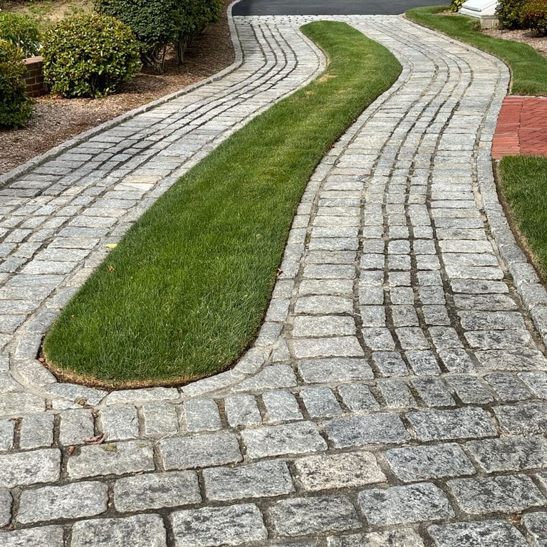 Application image for Gray Granite Cobblestone - Rectangles. The image shows this product being used in a driveway pavers application. The surface finish shown is tumbled. The dominant color shown is gray; with beige, brown, green and white secondaries. The color mix creates a cool feeling. Speckled and blotched color patterns. Contains some mica and mineral inclusions.