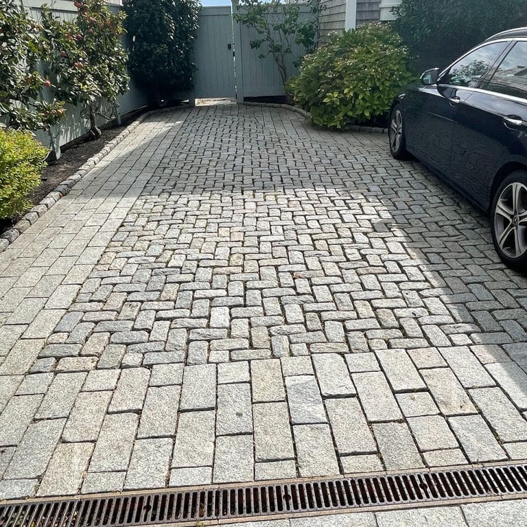 Application image for Granite Cobblestones - Gray. The image shows this product being used in a driveway pavers application. The dominant color shown is gray; with white and cream secondaries. Streaked and speckled color patterns. Contains some mica and mineral inclusions.