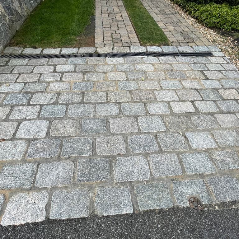 Application image for Granite Cobblestones - Gray. The image shows this product being used in a driveway pavers application. The surface finish shown is tumbled. Dominant colors shown are gray and white; with beige, blue, green and cream secondaries. The color mix creates a cool feeling. Streaked and speckled color patterns. Contains some mica and mineral inclusions.