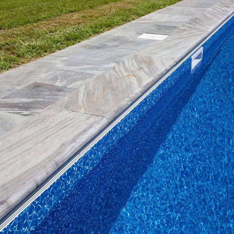 Best close-up image for Mystic Mountain Pool Coping. The image shows this product being used in a pool coping application. The surface finish shown is a combination of flamed and sawn. The dominant color shown is gray; with blue, green, purple and white secondaries. Exhibits "movement" and the color mix creates a cool feeling. Linear, streaked, veined and random color patterns. Contains mineral inclusions.