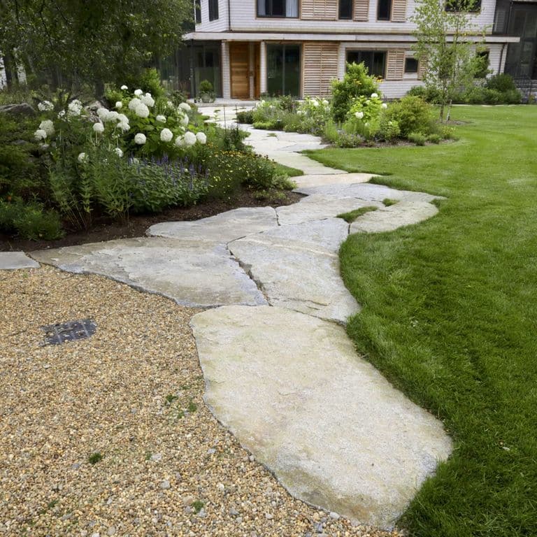 Application image for Dodlin Hill Irregular Flagstone. The image shows this product being used in a path pavers application. The surface finish shown is a combination of natural cleft and weathered. Dominant colors shown are beige, gray and cream; with white and tan secondaries. Exhibits "movement" and the color mix creates a warm feeling. Veined and speckled color patterns.