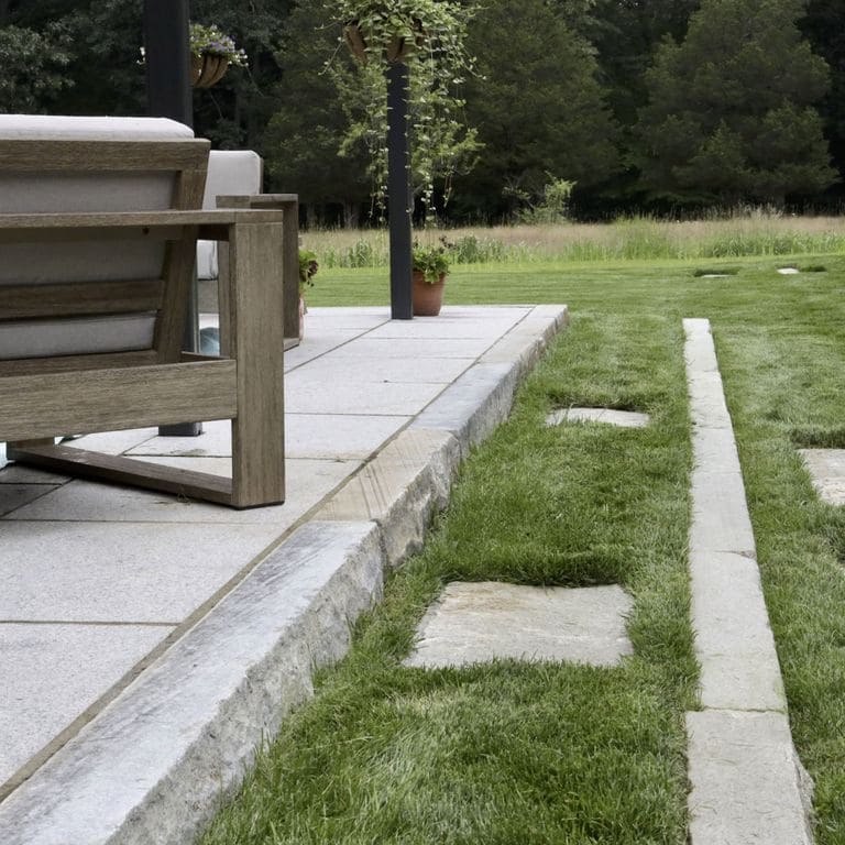 Application image for Reclaimed Third Generation Granite Curbstone Edging. The image shows this product being used for landscape curbing &amp; edging and monolithic steps. The surface finish shown is a combination of sawn, split-face with flat plane, weathered and worn. Dominant colors shown are gray and white; with beige and blue secondaries. The color mix creates a cool feeling. Fine speckled color pattern.