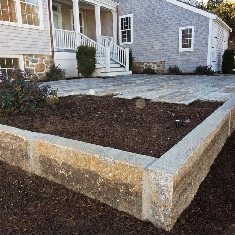 Application image for Reclaimed Third Generation Granite Curbstone Edging. The image shows this product being used in a landscape curbing &amp; edging application. The surface finish shown is a combination of sawn, split-face with flat plane, weathered and worn. Dominant colors shown are brown, gray and tan; with beige, orange/rust and white secondaries. The color mix creates a warm feeling. Speckled and blotched color patterns.