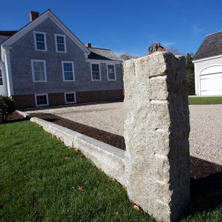 Application image for Reclaimed Third Generation Granite Curbstone Edging. The image shows this product being used in a landscape curbing &amp; edging application. The surface finish shown is a combination of sawn, split-face with flat plane, weathered and worn. Dominant colors shown are beige and gray; with white and cream secondaries. The color mix creates a cool feeling. Fine speckled and blotched color patterns.