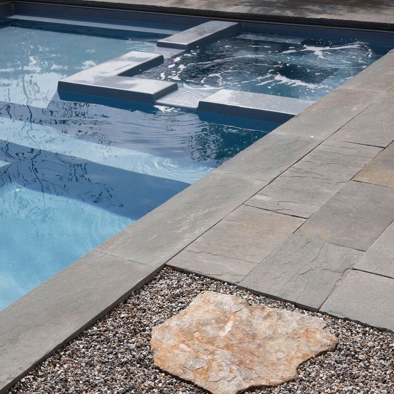 Feature image for Bluestone Natural Cleft Surface - Blue/Gray - Pool Coping. The image shows this product being used for pool coping, patio pavers and pool deck pavers. Dominant colors shown are blue and gray; with purple and tan secondaries. The color mix creates a cool feeling. Regular and cloudy color patterns.