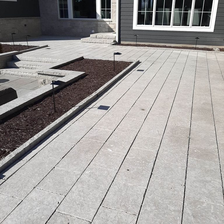 Application image for Madison Limestone - Pavers. The image shows this product being used for landscape curbing &amp; edging, patio pavers and monolithic steps. The surface finish shown is flamed. Dominant colors shown are beige and gray; with purple, white and cream secondaries. The color mix creates a cool feeling. Regular color pattern.