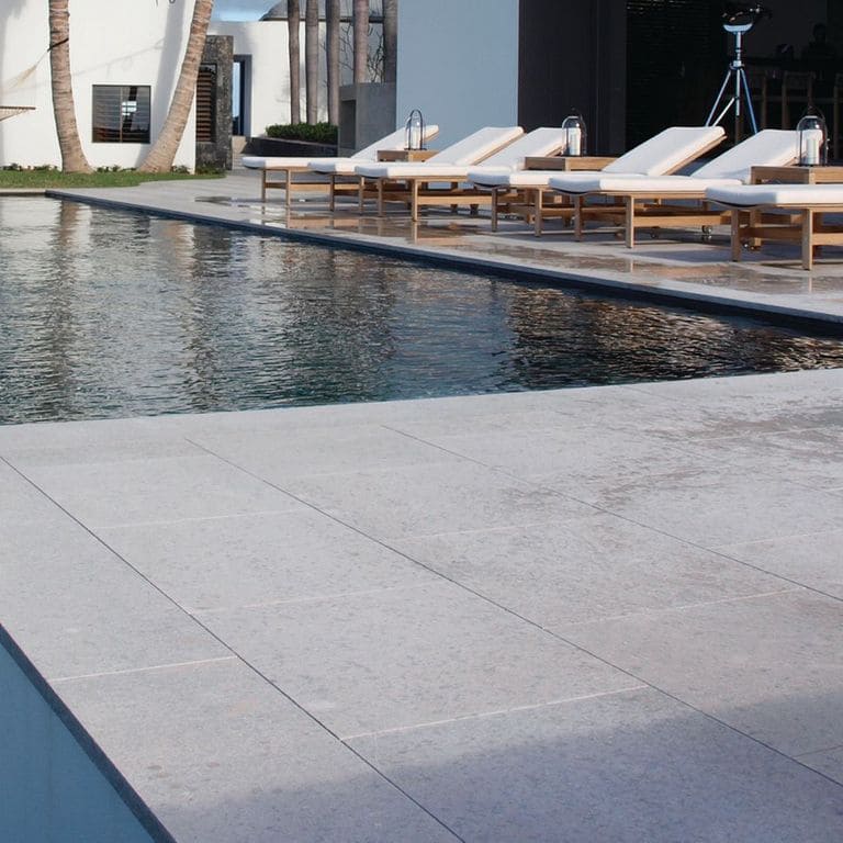 Best close-up image for Classimo Sky Limestone - Pool Deck. The image shows this product being used in a pool deck pavers application. The surface finish shown is sand blasted. Dominant colors shown are gray and white; with a blue secondary. The color mix creates a cool feeling. Regular and cloudy color patterns.