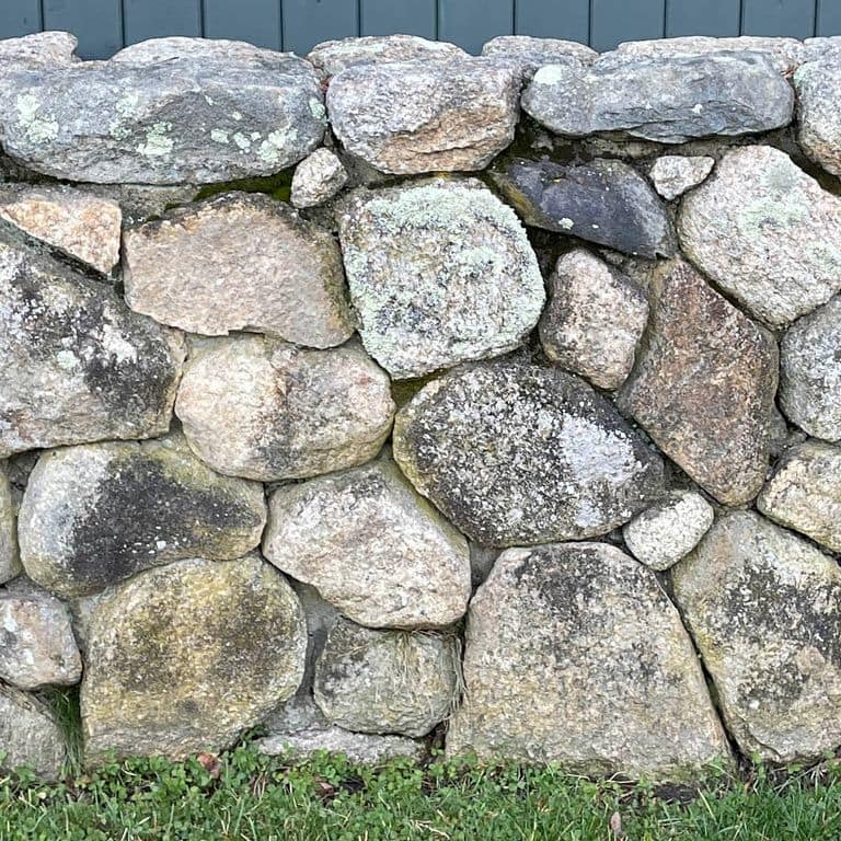 Best close-up image for New England Fieldstone Wallstone - Light. The image shows this product being used in a landscape wallstone application. The surface finish shown is a combination of natural and weathered. The dominant color shown is gray; with beige and cream secondaries. The color mix creates a warm feeling. Regular color pattern.