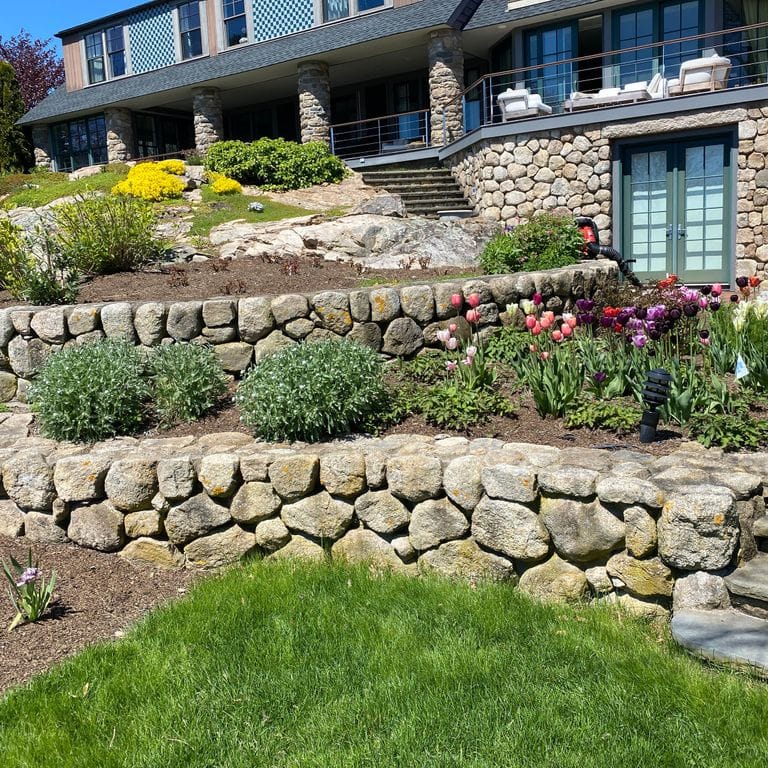 Feature image for New England Fieldstone Wallstone - Light. The image shows this product being used in a landscape wallstone application. The surface finish shown is a combination of natural and weathered. Dominant colors shown are beige and tan; with brown, orange/rust and cream secondaries. The color mix creates a warm feeling. Regular and blotched color patterns.