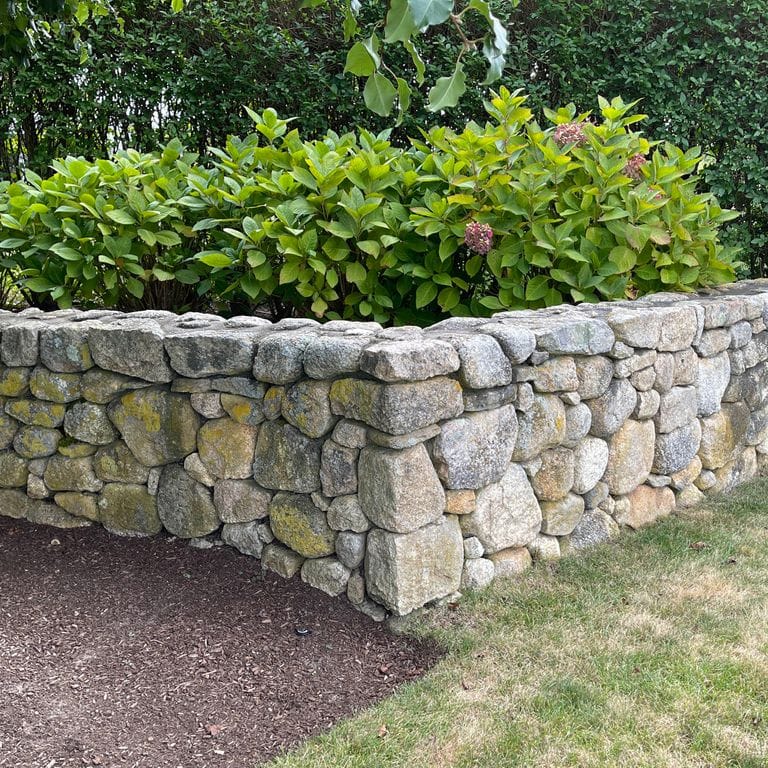 Application image for New England Fieldstone Wallstone - Light. The image shows this product being used in a landscape wallstone application. The surface finish shown is a combination of natural and weathered. The dominant color shown is gray; with beige, white, tan and cream secondaries. The color mix creates a warm feeling. Speckled and blotched color patterns.