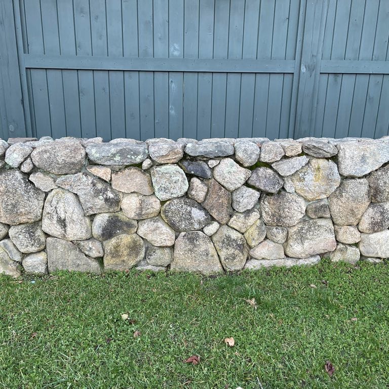 Application image for New England Fieldstone Wallstone - Light. The image shows this product being used in a landscape wallstone application. The surface finish shown is a combination of natural and weathered. The dominant color shown is gray; with beige, white, tan and cream secondaries. The color mix creates a warm feeling. Speckled, blotched and random color patterns.