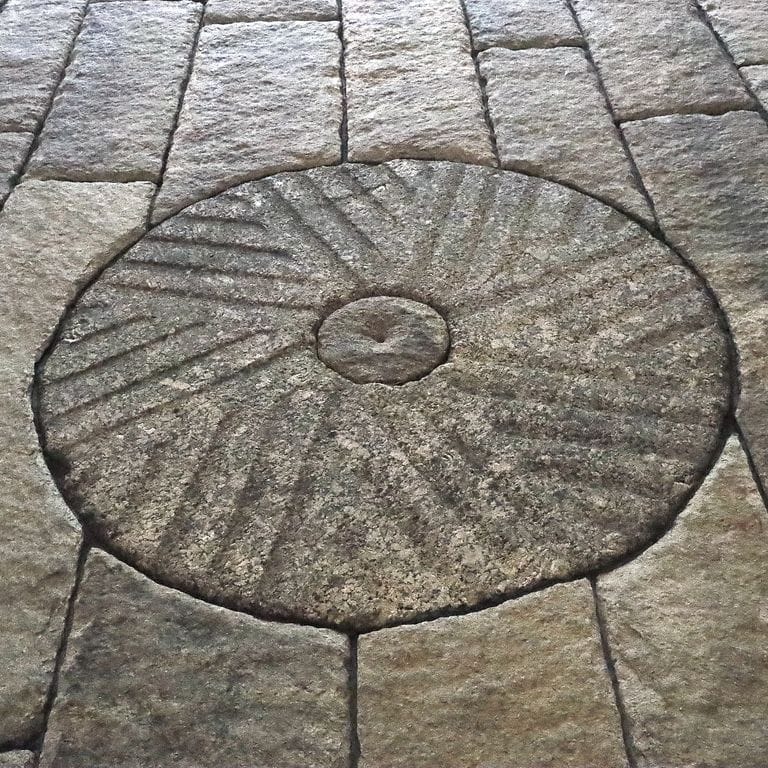 Best close-up image for Reclaimed Antique Mill Stones. The image shows this product being used in an interior floor application. The surface finish shown is a combination of carved, chiseled, weathered and worn. The dominant color shown is gray; with beige, black, brown, orange/rust and tan secondaries. The color mix creates a warm feeling. Speckled and blotched color patterns.