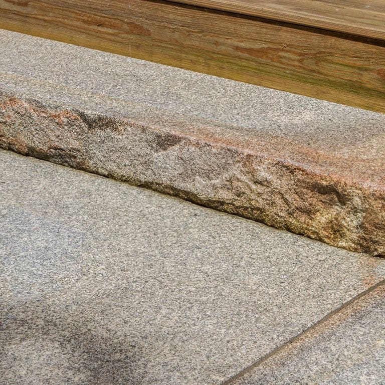 Application image for Cape Neddick Full Color Granite - Flamed Top Live Edge Steps. The image shows this product being used in a monolithic steps application. The surface finish shown is flamed. Dominant colors shown are beige and gray; with brown, orange/rust, white and tan secondaries. Exhibits "movement". Fine speckled and speckled color patterns.