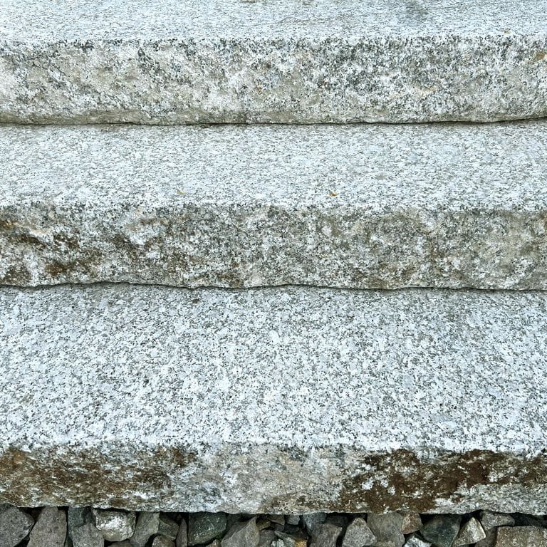 Best close-up image for Reclaimed Granite Bridge Blocks - Live Edge Steps. The image shows this product being used in a monolithic steps application. The surface finish shown is a combination of flamed, weathered and worn. Dominant colors shown are beige, blue and gray; with pink, purple, white, tan and cream secondaries. The color mix creates a cool feeling. Random and coarse speckled color patterns.
