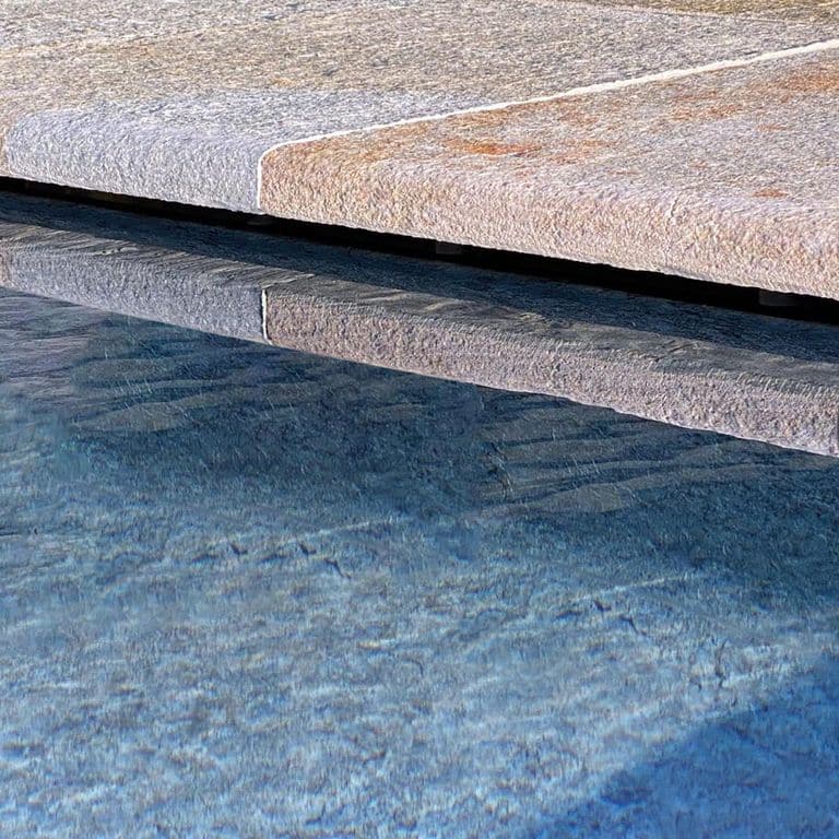 Best close-up image for Pietra Italia Full Color Gneiss - Pool Coping. The image shows this product being used in a pool coping application. The surface finish shown is natural cleft. Dominant colors shown are beige and gray; with beige, blue, brown, orange/rust, pink, purple, white, tan and cream secondaries. The color mix creates a warm feeling. Regular and fine speckled color patterns.