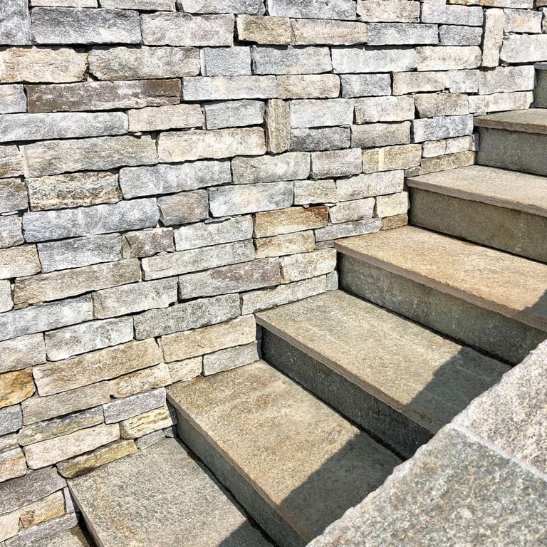 Best close-up image for Pietra Italia Full Color Gneiss - Treads. The image shows this product being used for wallstone cap and treads. The surface finish shown is natural cleft. Dominant colors shown are beige, gray and tan; with blue, brown, green, orange/rust, pink, purple, white and cream secondaries. The color mix creates a warm feeling. Regular, fine speckled and random color patterns. Contains some mica and mineral inclusions.