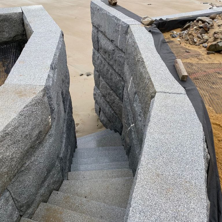 Application image for Granite - Dodlin Hill - Bridge Block. The image shows this product being used in a retaining wall application. The surface finish shown is natural cleft. The dominant color shown is gray. The color mix creates a warm feeling.