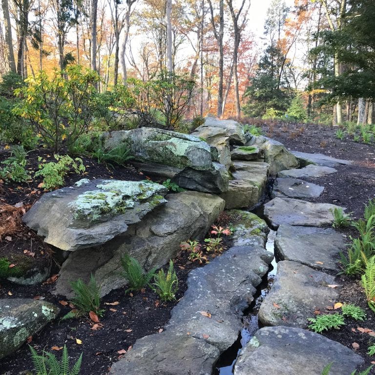 Application image for New England Fieldstone Boulders &amp; Ledgestone - Pool/Pond Coping. The image shows this product being used for water features and landscape boulders &amp; ledge. The surface finish shown is weathered. The dominant color shown is gray; with black, blue, green and white secondaries. The color mix creates a cool feeling. Regular and blotched color patterns.