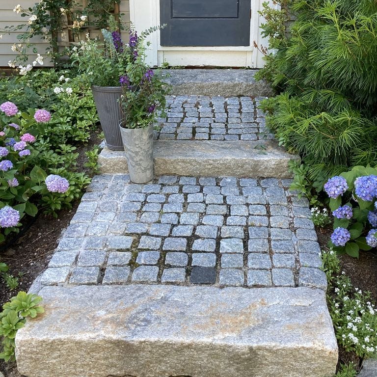 Feature image for Gray Granite Cobblestone - Squares. The image shows this product being used in a path pavers application. Dominant colors shown are blue, gray and white. The color mix creates a cool feeling. Speckled and coarse speckled color patterns. Contains some mica.